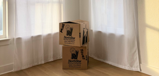 Two moving boxes are stacked in a corner of a new empty house. The boxes have NorthStar Moving logos and are labeled "mine" and "yours".